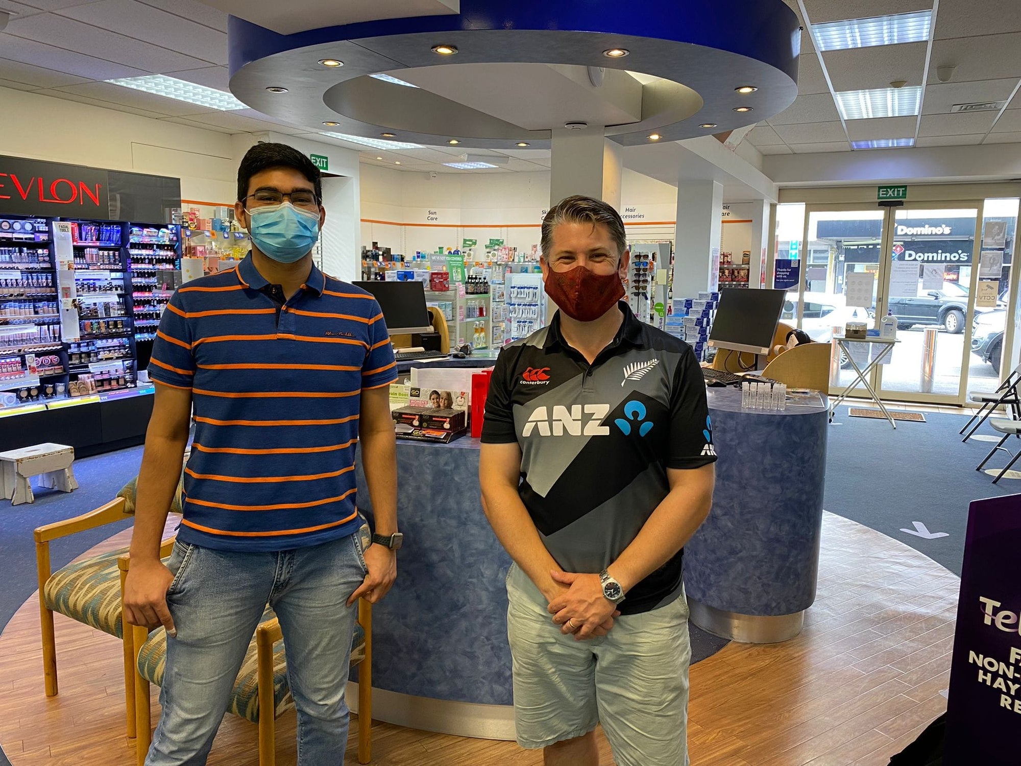 Two men standing inside the pharmacy store, wearing face masks, and smiling behind them.